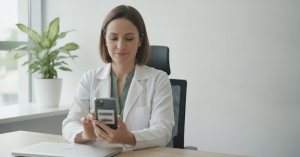 Marketing médico: médica de jaleco branco sentada à mesa do consultório olhando o celular, com notebook fechado e planta ao lado
